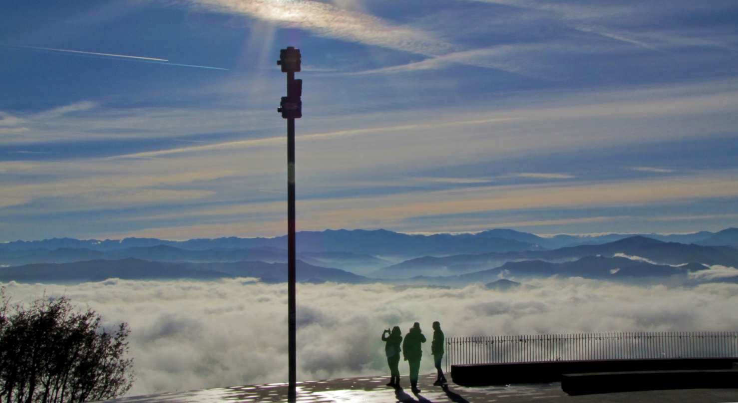 foto panorámica desde El Cristo. Se ve un mar de nubes sobre los montes del fondo. Tres personas contemplan el paisaje descrito.